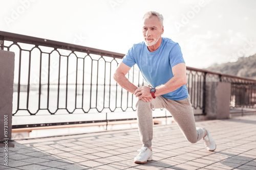 Sport forever. Senior confident unshaken making man exercises on the quay stretching legs and looking aside.