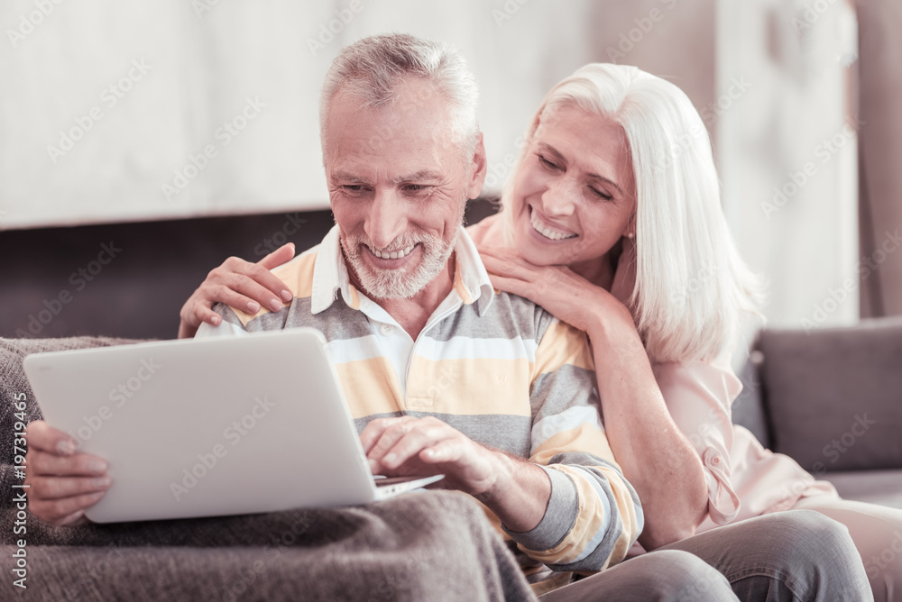 Funny moments. Interested aged pleasant couple spending time together at home smiling and working with the laptop.