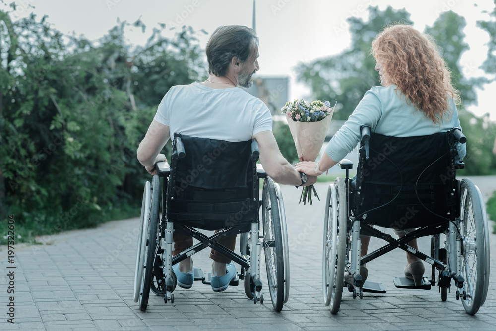 Warm atmosphere. Relaxed woman sitting in wheelchair and taking flowers while looking at her boyfriend