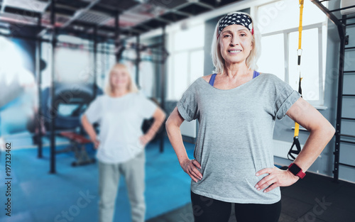 Get up and move. Hazel eyed elderly lady posing into the camera with her hands on the hips while standing in a gym and taking a group exercise class.