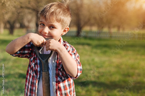 Like gardening. Cheerful little boy standing in the garden and holding shovel while looking at you
