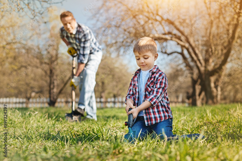 Enjoy working outdoors. Pleasant little boy digging the ground while his loving father standing in the background Enjoy working outdoors. Pleasant little boy digging the ground while his loving father standing in the background