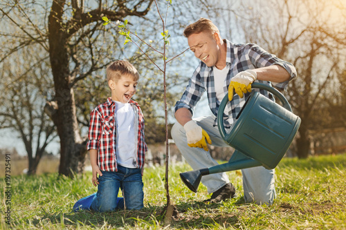 Eco friends. Positive father and his little son expressing joy and watering a tree while being involved in gardening