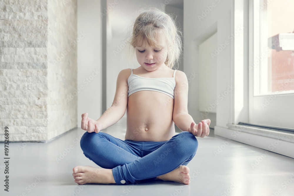 Little blond girl in white top and blue leggings doing a yoga exercise in the room