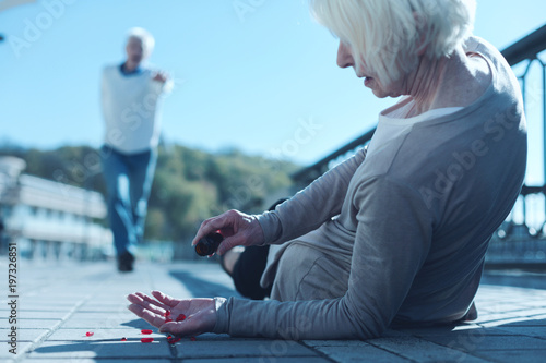 Results of missing my medication intake time. Selective focus on a poor woman lying on the ground and shaking a pill out of a bottle while trying to soothe her pain after falling down.