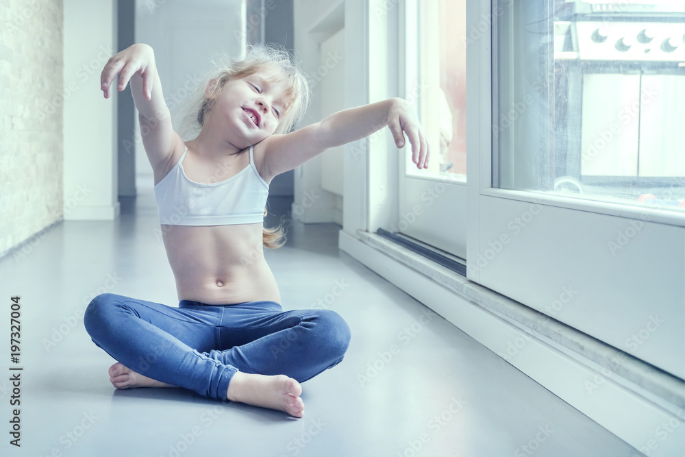 Little blond girl in white top and blue leggings doing a yoga exercise in the room