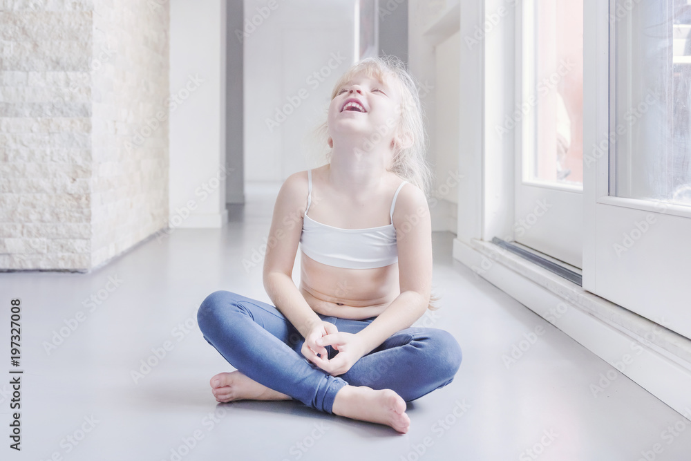 Happy little blond girl in white top and blue leggings sitting in a room  near the window Stock Photo | Adobe Stock