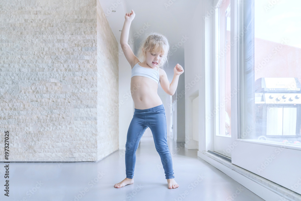 Little blond girl in white top and blue leggings dances in the room near the window Little blond girl in white top and blue leggings dances in the room near the window
