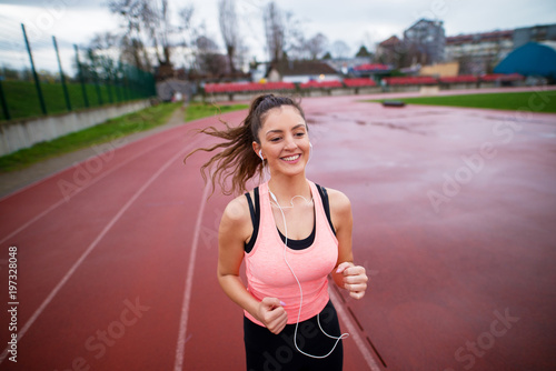 Portrait of attractive young happy fitness girl jogging while listening to music outside on running track.