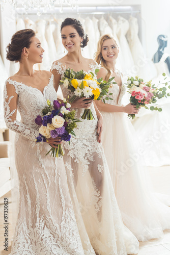 Smiling women in wedding dresses with flowers in wedding atelier