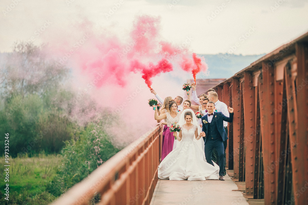 Newlyweds and their friends running with colors smoke bombs at old stone bridge. Happy wedding moments. Bride and groom with bridesmaids and groomsmen having fun at wedding day.