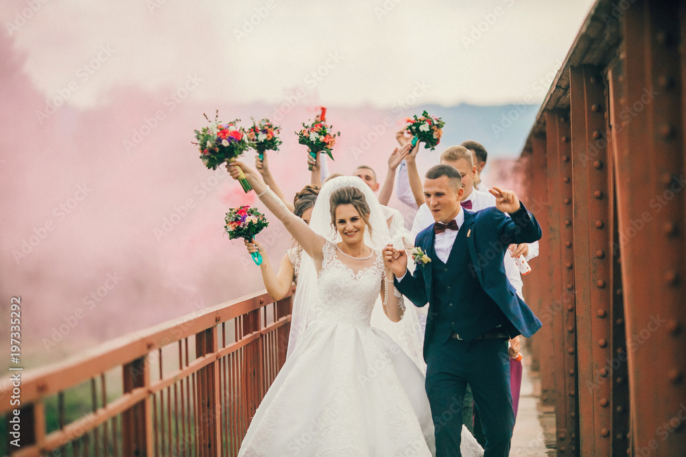Newlyweds and their friends running with colors smoke bombs at old stone bridge. Happy wedding moments. Bride and groom with bridesmaids and groomsmen having fun at wedding day.