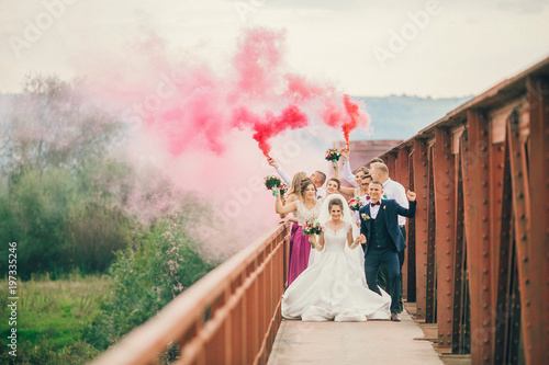 Obraz na płótnie Newlyweds and their friends running with colors smoke bombs at old stone bridge. Happy wedding moments. Bride and groom with bridesmaids and groomsmen having fun at wedding day. w salonie