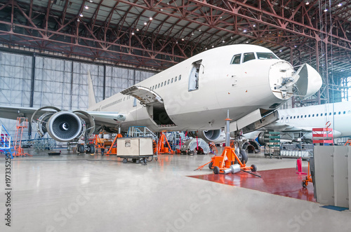 Passenger airplane on maintenance of engine and fuselage repair in airport hangar. Aircraft with open hood on the nose and engines, as well as the luggage compartment.