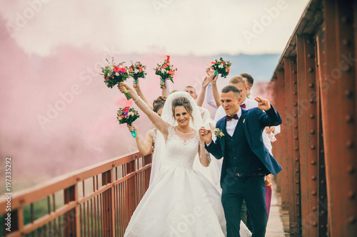 Obraz na płótnie Newlyweds and their friends running with colors smoke bombs at old stone bridge. Happy wedding moments. Bride and groom with bridesmaids and groomsmen having fun at wedding day. w salonie