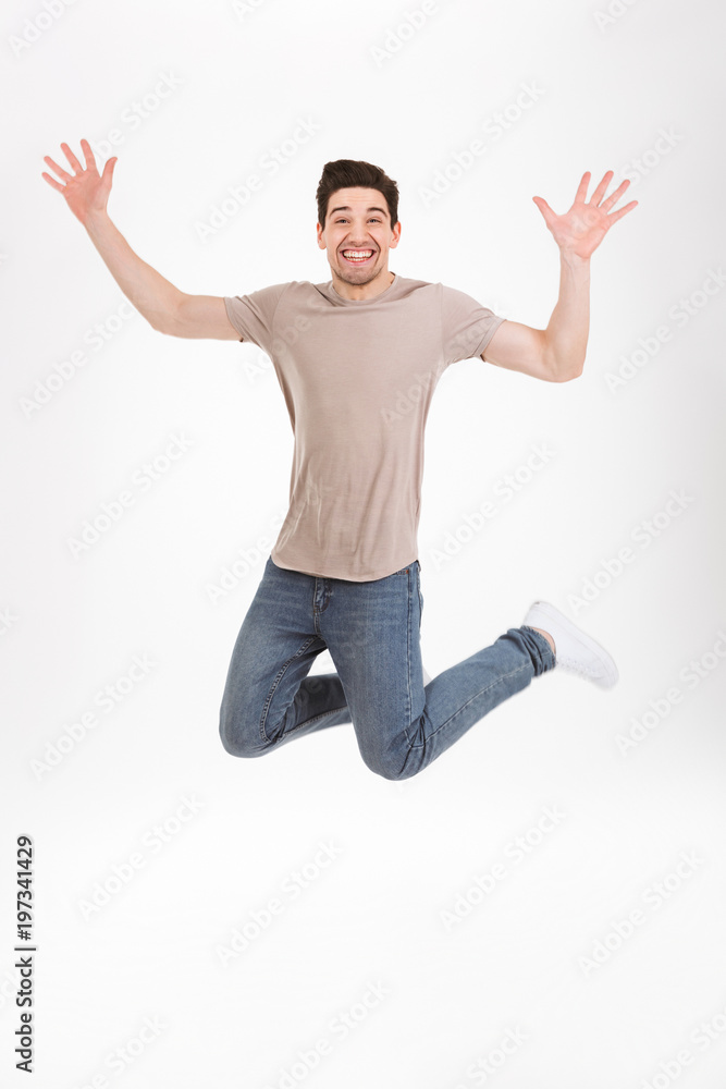 Full-length photo of excited man 30s in casual t-shirt and jeans levitating while expressing triumph and happiness throwing up hands, isolated over white background
