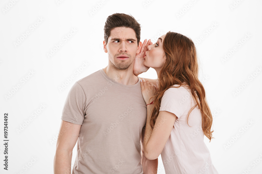 Photo of cute woman with beautiful long brown hair saying breathtaking rumors or news in ear of caucasian muscular man, isolated over white background