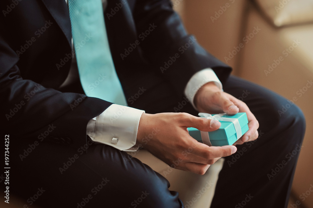 Handsome groom in a wedding suit waiting for the bride in a hotel room with present for grilfriend in his hands. Elegant businessman in a suit and tie. The groom suit Handsome groom in a wedding suit waiting for the bride in a hotel room with present for grilfriend in his hands. Elegant businessman in a suit and tie. The groom suit