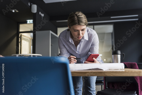 Young woman with cell phone and plan at table in office