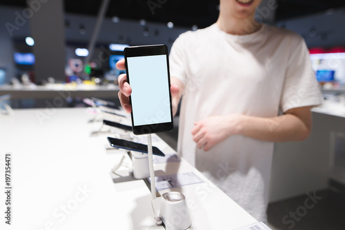 A man shows a smartphone with a white screen in the camera. Phone with a white screen on the background of the electronicsstore. Buy a phone in the store.