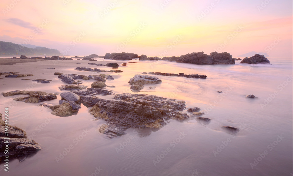 Sunrise scenery of a beautiful rocky beach on northern coast of Taiwan with an island on distant horizon under dramatic dawning sky & golden sunlight reflecting on the seawater (Long Exposure Effect)