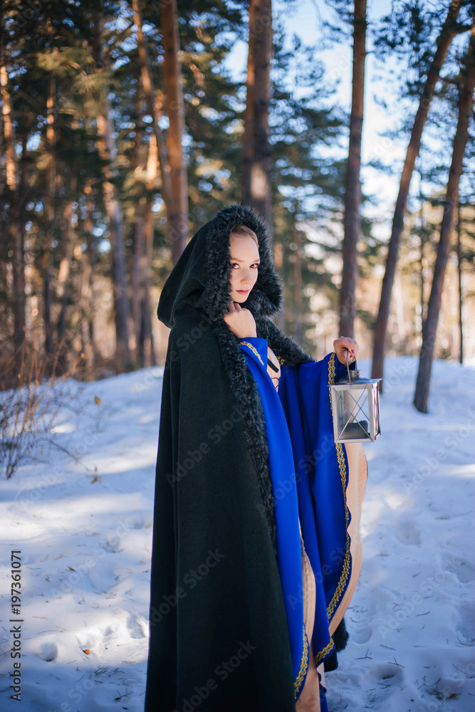 Beautiful girl in a medieval dress in blue and in a warm raincoat. A model with white hair in a winter forest with a large lamp. Beautiful girl in a medieval dress in blue and in a warm raincoat. A model with white hair in a winter forest with a large lamp.