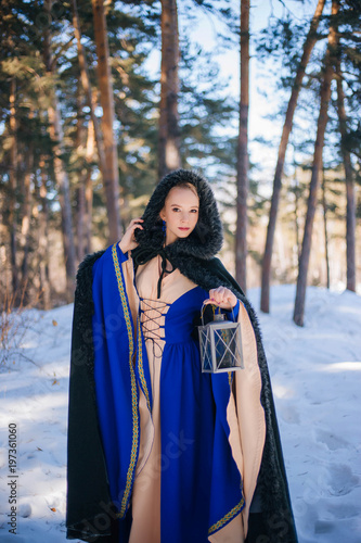 Beautiful girl in a medieval dress in blue and in a warm raincoat. A model with white hair in a winter forest with a large lamp.