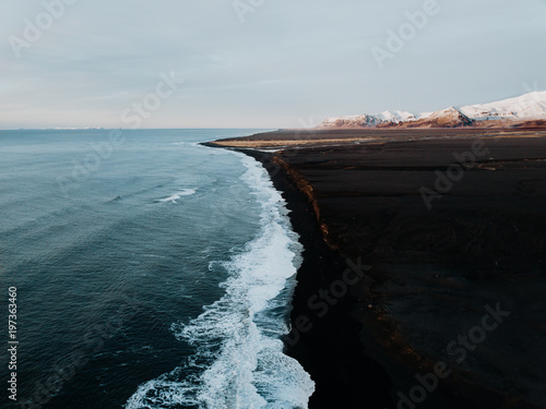 The coast of Iceland with a black sand beach and some mountains in the background, photographed from a drone