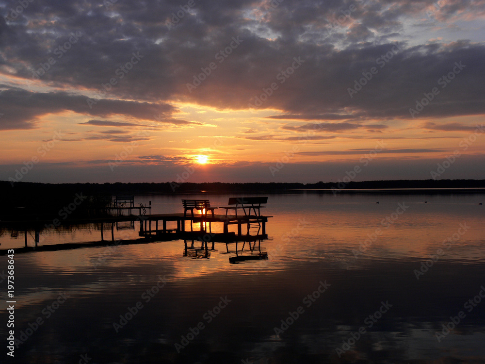hölzerner Steg mit Sitzbänken vor einem stimmungsvollen Sonnenuntergang am Arendsee mit dramatischen Wolken und spiegelglattem Wasser