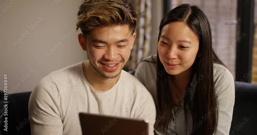 4K Happy Asian couple at home looking at digital tablet & laughing 4K Happy Asian couple at home looking at digital tablet & laughing