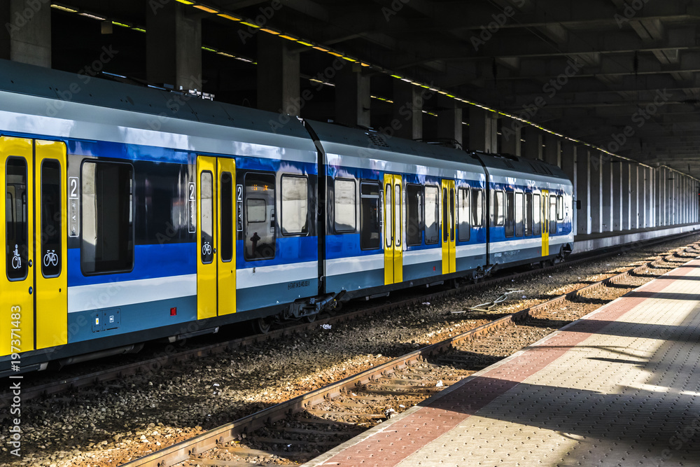 Fototapeta premium Railway station with train and platform. Covered old railway station with a modern blue and yellow train at sunny day.