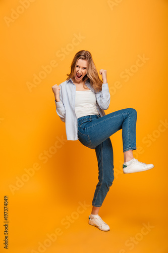Full length picture of pleased pretty woman yelling and clenching fists, acting like winner or lucky person, isolated over yellow background