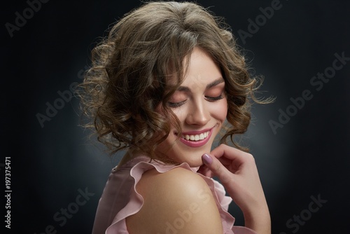 Studio portrait of a nice young girl with brown curly hair and light day make up. Model is looking down and smiling. Woman feels happy and touching her face. Backview on dark grey background. photo