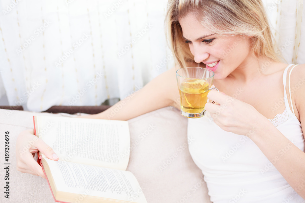 Beautiful happy young woman reading a book and drinking tea while sitting in pajamas in a brightly lit room. High angle view. Beautiful happy young woman reading a book and drinking tea while sitting in pajamas in a brightly lit room. High angle view.