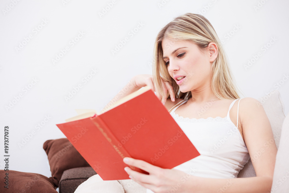 Beautiful happy young woman reading a book. Sitting in pajamas on a sofa in a brightly lit room Beautiful happy young woman reading a book. Sitting in pajamas on a sofa in a brightly lit room
