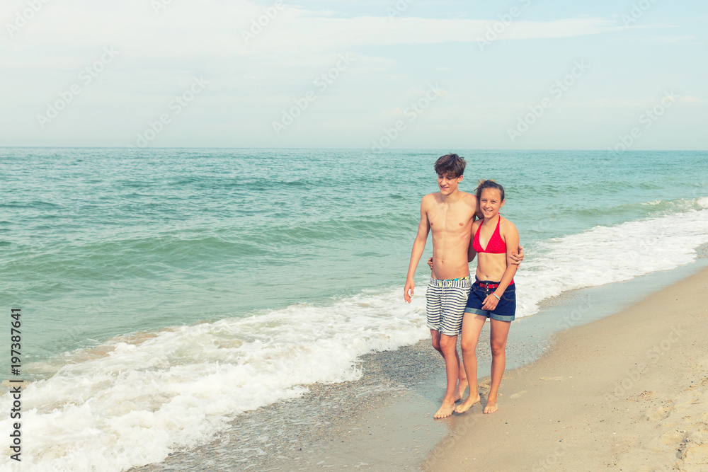 Two teenagers: a girl and a boy with blond hair, dressed in a swimsuit walks returning to the viewer on the sea beach.