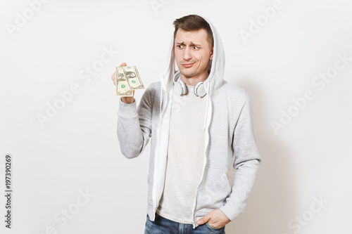 Young handsome male student in t-shirt, blue jeans and light sweatshirt with hood with headphones holds two one-dollar bills and perplexedly looks isolated on white background. Concept of emotions photo