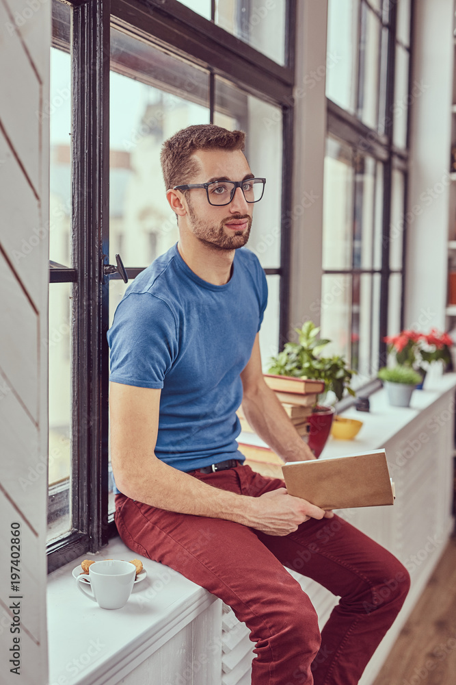 Young caucasian student reading a book sitting on a window sill. Young caucasian student reading a book sitting on a window sill.