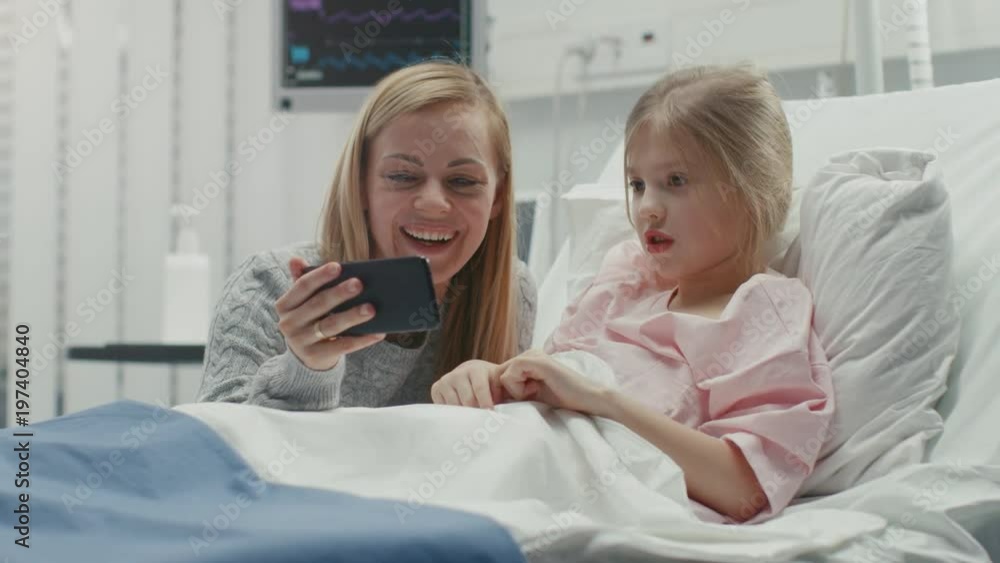 Cute Little Girl Lies on a Bed in the Children's Hospital, Her Mother Sits Beside, They Watch Cartoons/ Funny Videos on Smartphone. Modern Pediatric Ward. Shot on RED EPIC-W 8K Helium Cinema Camera. Cute Little Girl Lies on a Bed in the Children's Hospital, Her Mother Sits Beside, They Watch Cartoons/ Funny Videos on Smartphone. Modern Pediatric Ward. Shot on RED EPIC-W 8K Helium Cinema Camera.