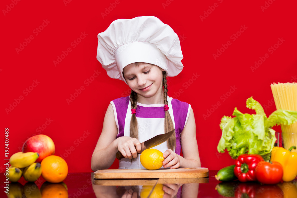 Happy little girl in chef uniform cuts fruit in kitchen. Kid chef. Cooking Process Concept Happy little girl in chef uniform cuts fruit in kitchen. Kid chef. Cooking Process Concept
