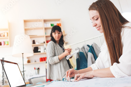 Team of two women dressmaster create new concept clothing with contemporary laptop in sewing showroom. Small business. Seamstress working with cloth in workshop
