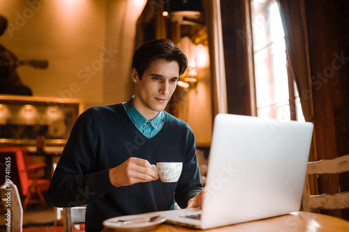 Young businessman holding mug of coffee while working on laptop computer in coffee shop, male freelancer writing text on laptop keyboard while enjoying cup of cappuccino in modern cafe