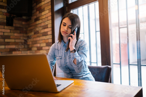 Gorgeous female speaking on mobile phone with smile while sitting at wooden table with open laptop computer, young student or businesswoman at work break with net-book in modern coffee shop interior.