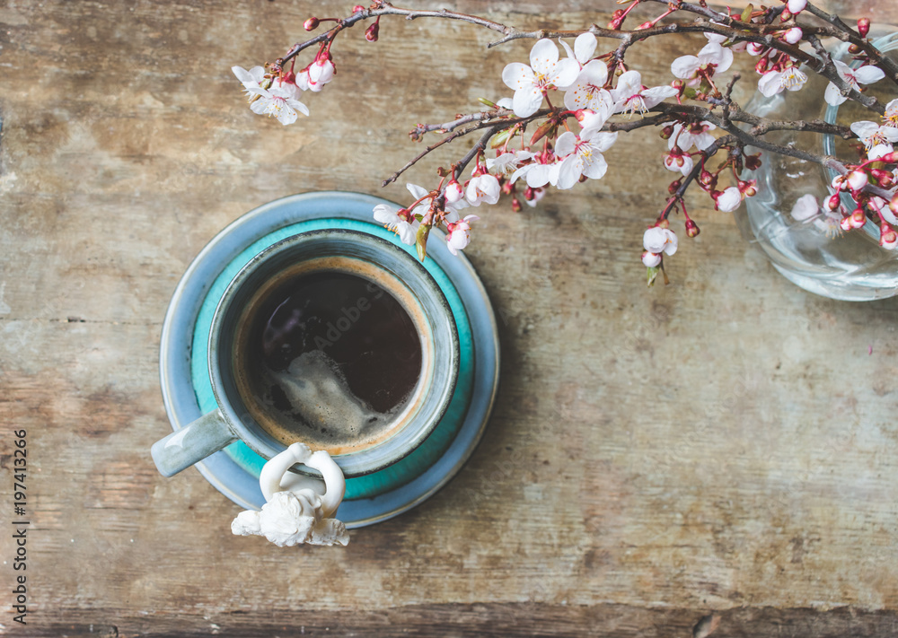 Close up view of a white angel figurine on a cup of coffee and fresh spring tree branches blooming with pink and white flowers on wooden background. Hello spring or Happy Easter background