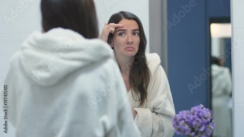 Frustrated young woman standing in front of the bathroom mirror in the morning and looking at her skin, hair and teeth. Upset woman checking acne and dull dry thin hair dissatisfied with her look.