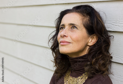 Fascinating mature woman with optimistic look leans on white wood plank wall background. Middle aged lady with hopeful, positive expression. Plan, imagination, creative concept