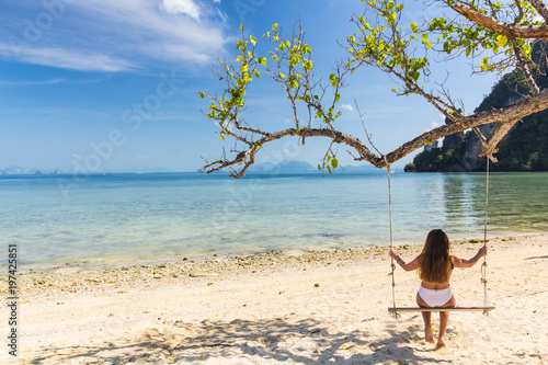 Tourist girl in bikini sitting on wooden swing On the white beach, the clear blue sky.