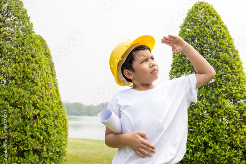 Asian boy in white t-shirt with yellow helmet dress like engineer raise his arm on head level with garden scene