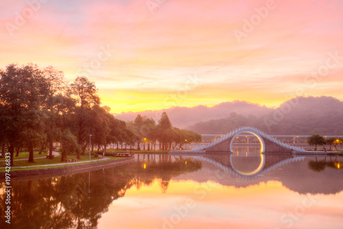 Scenery of an urban park lit up by golden sunlight with reflections of an arch bridge and glowing sky on the smooth lake water ~ Early morning scene of Dahu Community Park in Nahu,Taipei City Taiwan photo