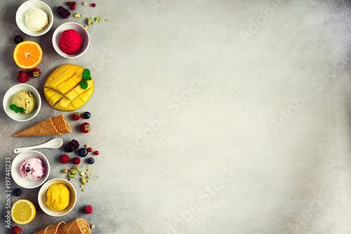 Ice cream balls in bowls, waffle cones, berries, orange, mango, pistachio on grey concrete background. Colorful collection, flat lay, summer concept, top view